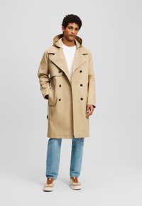 Gabardina beige con un diseño de doble botonadura, gran cuello y bolsillos laterales. Combinada con jeans de color azul claro y zapatillas beige.