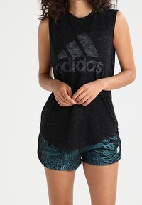 Musta hihaton paita, jossa on suuri harmaa adidas-logo, yhdistettynä teal-värisiinShortseihin, joissa on vihreä lehtikuosi ja tiivis muotoilu.