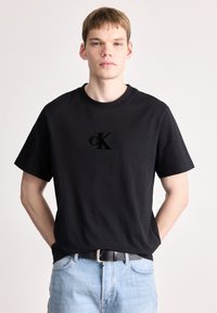 Melna kokvilnas T-krekls ar īsām piedurknēm, ar teksturētu melnu "CK" logotipu uz priekšpuses. Ar apaļu kakla izgriezumu, apvienots ar gaiši ziliem džinsiem.