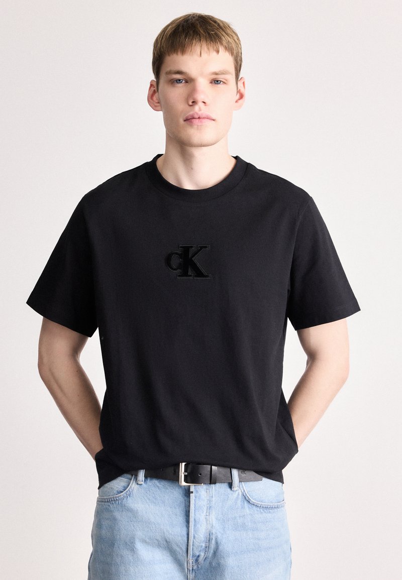 Melna kokvilnas T-krekls ar īsām piedurknēm, ar teksturētu melnu "CK" logotipu uz priekšpuses. Ar apaļu kakla izgriezumu, apvienots ar gaiši ziliem džinsiem.