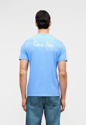 Homme portant un t-shirt Calvin Klein bleu clair et un jean bleu, vu de dos, avec un logo blanc en haut du dos.