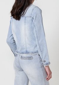 Light blue denim jacket with braided accents at the hem and cuffs, featuring a classic cut and front pockets, shown from the back.