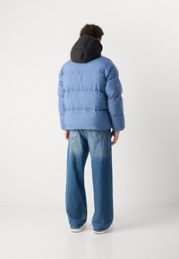 Light blue puffer jacket with black hood, featuring a quilted design. Paired with faded wide-leg jeans and white sneakers, viewed from the back.