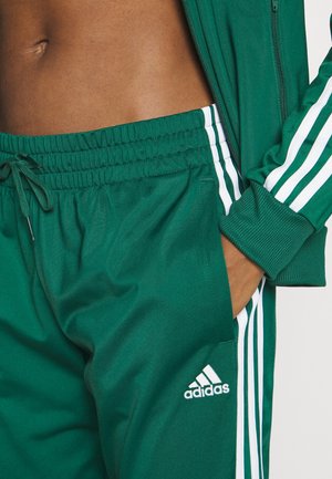 Pantalon de survêtement et veste Adidas verts avec des rayures blanches portés par une personne avec une main dans la poche, montrant le ventre découvert.