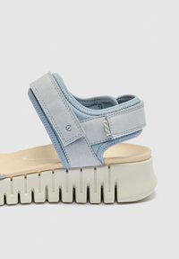 Ανοιχτό-μπλε πέδιλο με ανοιχτά δάχτυλα, ρυθμιζόμενα λουριά και χοντρή, τμηματοποιημένη σόλα εκρού, εμφανίζεται από το πλάι πάνω σε λευκό φόντο.