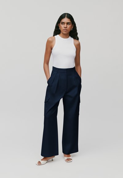Haut sans manches blanc associé à un pantalon cargo large bleu marine avec des plis à l'avant et des poches latérales ; le mannequin se tient sur un fond neutre.