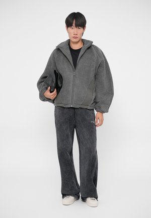 Harmaa fleece-takki, jossa on vetoketju ja ylisuuret hihat, yhdistettynä löysiin tummanharmaisiin housuihin. Takissa on tukeva tumma runko ja teksturoitu kangas.