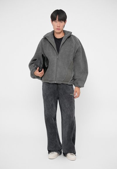Veste en polaire grise avec un zip sur le devant et des manches surdimensionnées, associée à un pantalon noir gris ample. La veste possède un corps uni de couleur sombre et un tissu texturé.