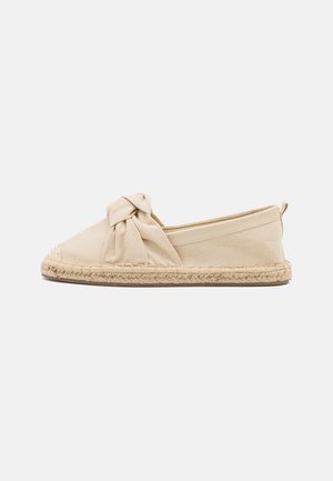 Beige lærred slip-on sko med en knyttet stofdetalje på toppen og fletbunden jutesål, vist fra siden på en hvid baggrund.