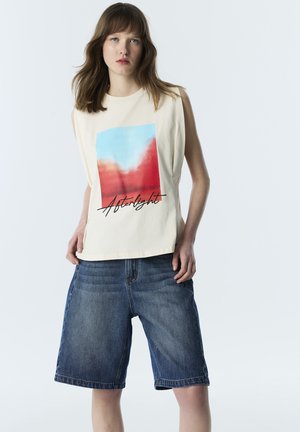 Kobieta z brązowymi włosami do ramion, ubrana w kremowy top bez rękawów z czerwono-niebieską grafiką i napisem „Afterlight”, w połączeniu z dżinsowymi szortami do kolan.