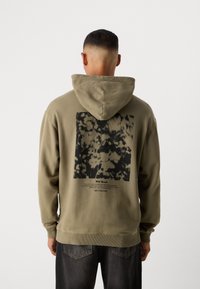 Man met kort haar, gekleed in een olijfgroene hoodie met zwart abstract bloemenpatroon en tekst op de rug, gecombineerd met zwarte spijkerbroek.