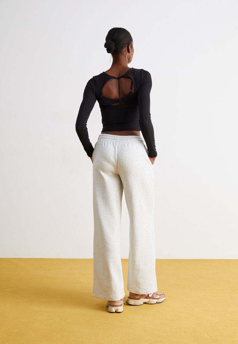 Vrouw met een haar in een knot draagt een zwart long-sleeve top met een uitsnede op de rug, grijze wijde sweatpants en roze sneakers op een gele vloer.