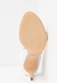Semelle d'une chaussure à talon haut blanche avec une semelle inférieure en cuir lisse beige et une pointe de talon étroite, inclinée sur un fond blanc.