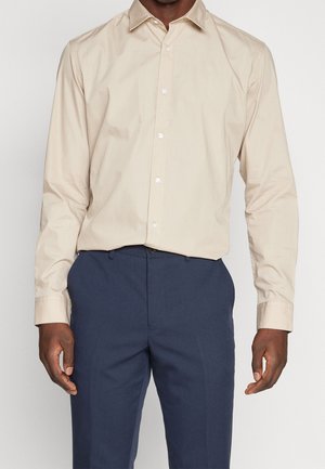 Homme portant une chemise beige à manches longues boutonnée, rentrée dans un pantalon habillé bleu marine, debout devant un fond clair uni.