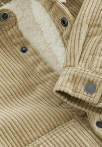 Gros plan sur une veste en velours côtelé beige côtelée avec des boutons noirs et une doublure intérieure en polaire blanche douce visible.