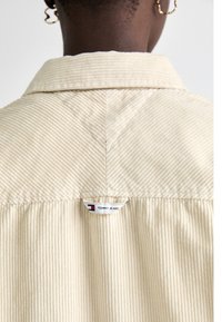 Camisa de pana color crema con cuello, que presenta un ribete vertical y una etiqueta con el logo en la parte posterior del cuello. Tejido texturizado y corte relajado.