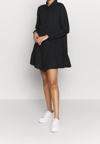 Abito camicia nero, lungo fino al ginocchio, con maniche lunghe, davanti a bottoni e orlo arricciato. Abbinato a sneakers bianche. Tessuto morbido.