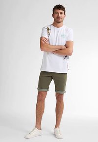 Bílé bavlněné tričko s grafikou palmy, olivově zelené šortky s ohrnutými lemy a bílé tenisky. Model stojí se založenýma rukama.