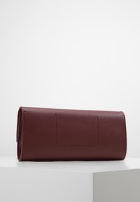 Tmavě hnědá kožená kabelka leží na bílé podložce, s viditelnými stehy a víkem na překlápění.