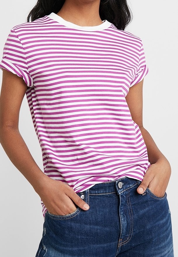 Osoba v růžovém a bílém pruhovaném tričku s krátkým rukávem, zastrčeném do tmavě modrých džínů, s rukama v kapsách.