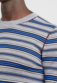 Camiseta de rayas con base gris y rayas azules, negras y blancas, mangas cortas y un escote redondo con un acento burdeos en el hombro.