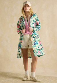 Bloemenregenjas met groene, roze en blauwe patronen; roze shirt; beige shorts; witte sokken; beige schoenen; ontspannen pasvorm, knielang.