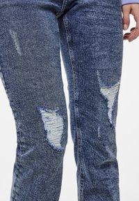 Denim jeans in dark blue with distressed patches. Features frayed holes near the knees and a textured fabric with a worn look.