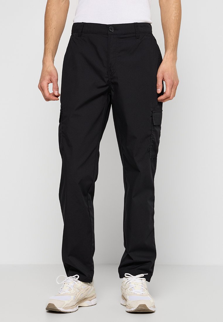 Homme portant un pantalon cargo noir avec des poches latérales et des baskets blanc cassé, se tenant devant un fond blanc uni.