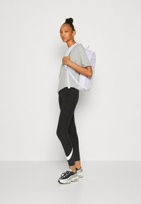 Grå kortärmad tröja, svarta leggings med vit sidostripe, vit ryggsäck och svarta sneakers med silverdetaljer, stående mot en enhetlig bakgrund.