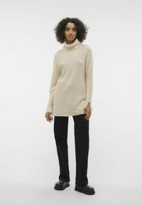 Beige gebreide coltrui met lange mouwen, zijsplitten, gecombineerd met zwarte wijde broeken en chunky zwarte schoenen.