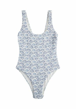 Donna con un costume da bagno blu e bianco con motivo a conchiglie, in piedi su una spiaggia sabbiosa vicino a delle rocce e un asciugamano rosso a righe.