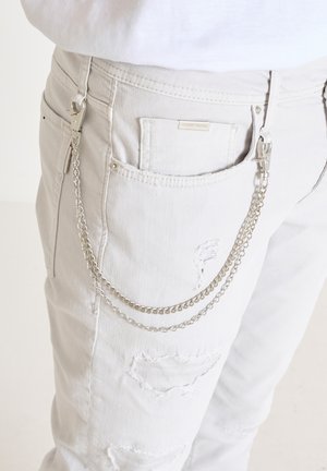 White denim jeans feature distressed patches and a silver chain detail attached to the back pocket with a clip.