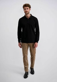 Man in een zwart langemouwen polo shirt, beige broek en zwarte schoenen die tegen een effen lichte achtergrond staat.