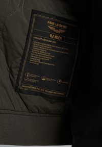 Lekki pilot jacket w matowym oliwkowym kolorze z pikowanym wykończeniem. Posiada czarną metkę ze złotymi detalami i akcentami. Wykonany z materiałów recyklingowych.
