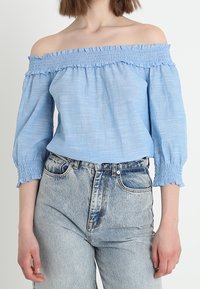 Ljusblå avaxlad blus med rynkad elastisk halsringning och strukturerad tyg, kombinerad med högmidjade blekta jeans.