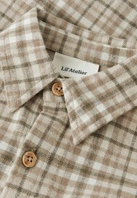 Pruun ja beež ruuduline nööbiga särk, millel on krae ja puidust nupp, kangasilt „Lil'Atelier.”
