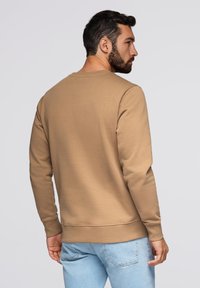 Felpa beige con maniche lunghe e collo a giro, caratterizzata da polsini e orlo a coste. Indossata con jeans azzurri chiari. Tessuto liscio e textured.
