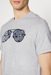 Camiseta gris de manga corta con un gran gráfico de gafas de sol de gran tamaño en azul marino, con el nombre "MICHAEL KORS" en un patrón repetido.