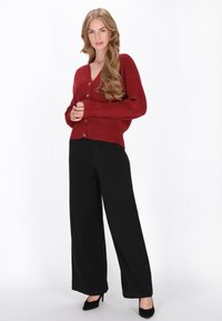 Piros kötött kardigán mély V-alakú nyakkal és gombos elülső részével, fekete, bő szabású nadrággal és fekete magassarkú cipőkkel párosítva. Simább textúra.