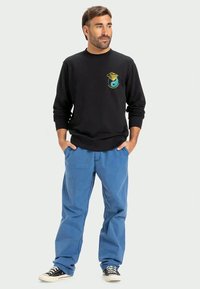 Sweatshirt preto com gráfico colorido, calças azuis e ténis pretos. Ajuste casual com punhos e bainha canelados. Design simples, sem padrões.