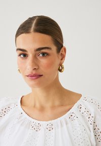 White blouse with eyelet detailing, round neckline, and soft texture, paired with large gold hoop earrings. Minimal makeup, neutral background.