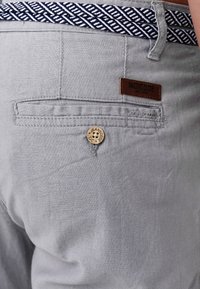 Grå byxor med strukturerad yta, med midjeband i marinblått mönster, en träknapp med varumärke och en läderlapp med logotyp.