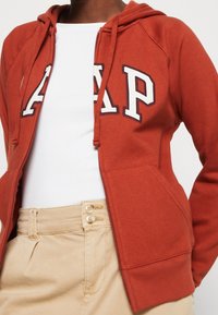 Persoon in een rode zip-up hoodie met het "GAP" logo, een witte shirt eronder, en beige hoge taillebroek met dubbele knopen.