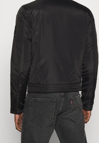 Giacca bomber nera in nylon con una texture liscia, vita e polsini a costine, con un'etichetta del logo sulla manica sinistra.