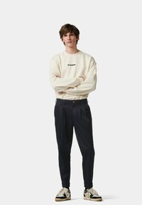 Lichtbeige sweatshirt met zwart logo, marineblauwe geplooide broek en zwart-witte sneakers met gouden accenten en textuurlinten.