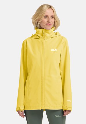 Blonde Frau trägt eine leuchtend gelbe wasserdichte Jacke mit hohem Kragen und Logos auf Brust und Ärmel, steht vor weißem Hintergrund.