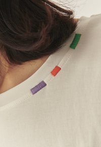 Biała koszulka z okrągłym dekoltem, ozdobiona kolorowymi haftowanymi akcentami w kolorach zielonym, pomarańczowym, fioletowym i niebieskim na kołnierzu. Gładka faktura.