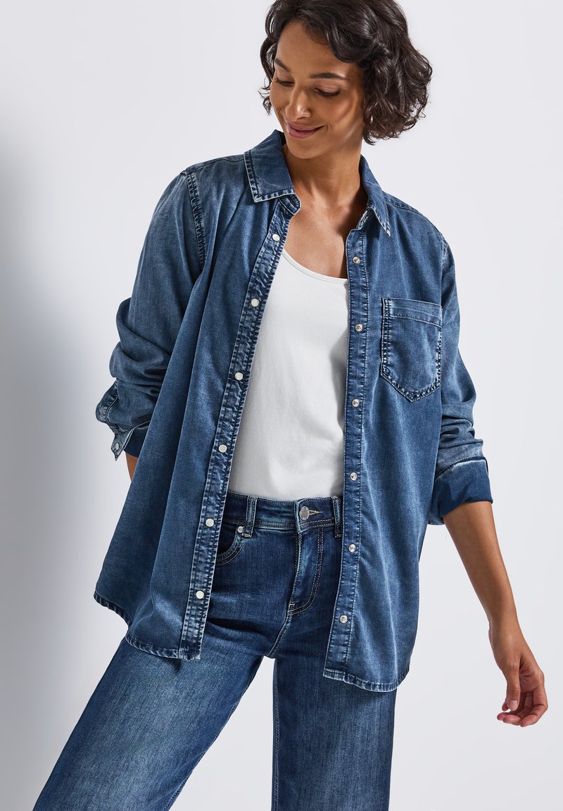 Dunkelblaue Jeanshemd mit Knopfleiste, Brusttasche und hochgekrempelten Ärmeln. Kombiniert mit blauen Jeans und einem weißen Tanktop.