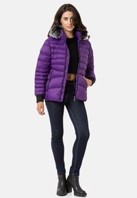 Lila Puffjacke mit horizontalen Nähten, Kunstpelzbesatz an der Kapuze, Reißverschlusstaschen und gerippten Bündchen. Getragen mit schwarzem Oberteil und dunkler Jeans.