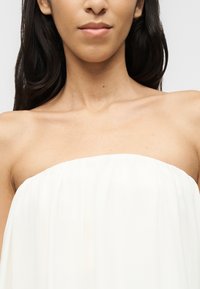 Γυναίκα με μακριά σκούρα μαλλιά που φοράει λευκό τοπ χωρίς τιράντες, εμφανίζοντας τους ώμους, τον λαιμό και το κατώτερο μέρος του προσώπου της, σε απλό φόντο.